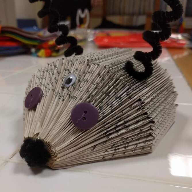 A book folded down to resemble a hedgehog. The book has two brown pipe cleaners as curled antennae, two purple button eyes, a wiggle eye centered on its face, and a brown pom pom nose.